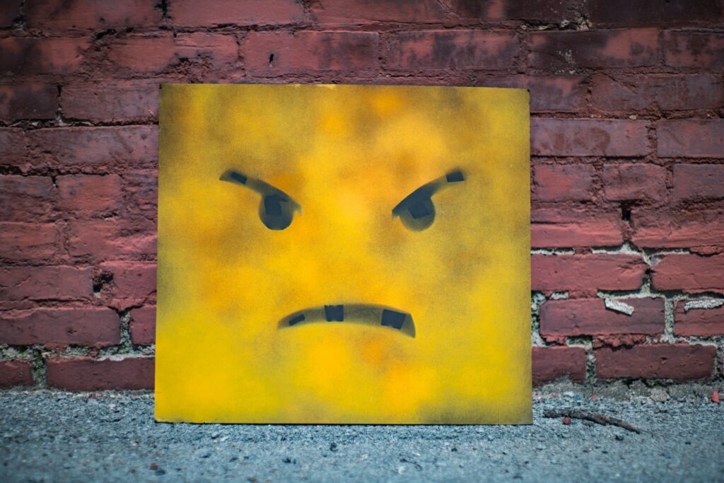 Eine rechteckige Metalltafel, die mit einem gelben grimmig dreinblickenden Smiley bemalt ist, lehnt an einer roten Ziegelwand.