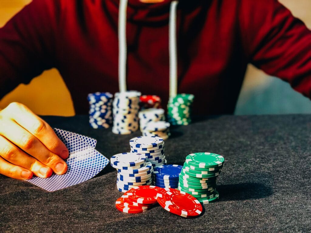 Ein Mann im roten Pullover sitzt an einem mit grauem Stoff überzogenen Tisch vor Jetons und hält einige Spielkarten in seiner rechten Hand.