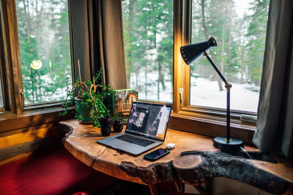 Auf einem rustikalen Holztisch steht ein geöffneter Laptop. Daneben liegt ein Smartphone. Der Blick nach draußen ist auf einen verschneiten Wald gerichtet.